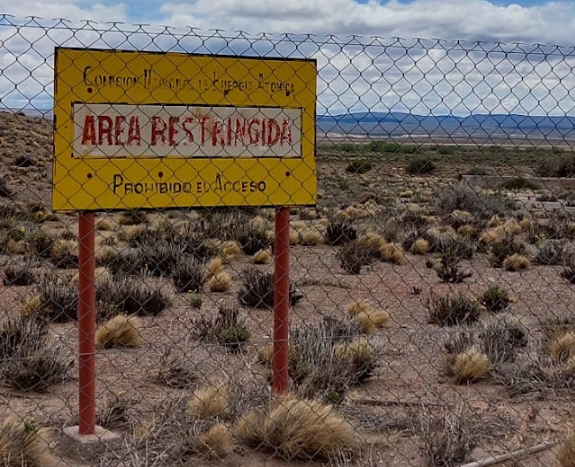 Alambrado y cartel. La remediación del predio de la antigua mina de uranio Los Adobes (Distrito Pichiñan, Chubut), que debe realizar la Comisión Nacional de Energía Atómica (el organismo que explotó la mina durante tres años), por ahora no pasa de eso. El pasivo ambiental de 85.000 toneladas de residuos contaminantes de Los Adobes (mayormente colas de mineral) es una pequeña fracción de las 6.000.000 de toneladas de residuos a lo largo y ancho del país. Fuente: https://huelladelsur.ar/2021/12/16/el-legado-radiactivo-del-distrito-uranifero-pichinan/
